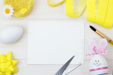White notepad on a white wooden background. The Happy Easter Bunny is created from an egg. The moment of creating an Easter decoration. top view, flat lay