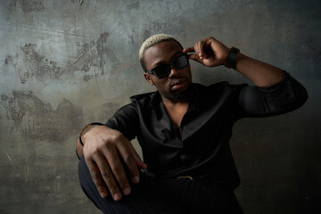 Stylish african handsome male model wearing black silk shirt, trousers and sunglasses, sitting leaning on grey concrete wall