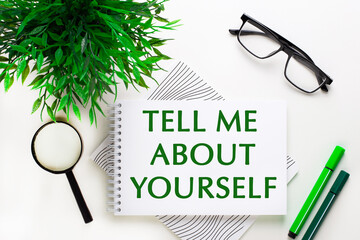 On a white background lies a notebook with words TELL ME ABOUT YOURSELF, glasses, a magnifying glass, green markers and a green plant