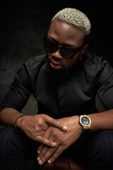 Elegant stylish handsome african man in black silk shirt, pants and sunglasses sitting with crossed hands in dark interior