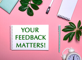 YOUR FEEDBACK MATTERS is written in green on a white notepad on a pink background surrounded by notepads, pens, white alarm clock and green leaves. Educational concept