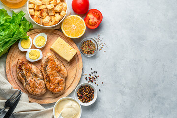 Products for making a traditional Caesar salad. Juicy chicken breast, eggs, cheese, sauce and herbs on a gray background.