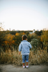 Naklejka na ściany i meble child stands with his back in the park. boy in denim clothes stands in the forest at sunset. child walks in the woods. stylish kid outdoors. child lost in the woods 