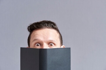 Young man staring in surprise at the camera peering out from behind an open book