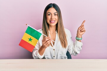 Young hispanic woman holding senegal flag sitting on the table smiling happy pointing with hand and...