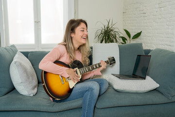 Young woman learning online how to play guitar following video tutorial on laptop at home