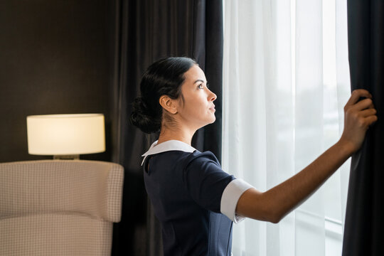 Side view of young chambermaid in uniform opening dark curtains on large window of hotel room before cleaning it in the morning