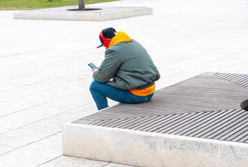 Mature man with headphones sitting on bench using phone. Full length portrait of caucasian  modern young man sitting and looking at mobile phone in the street.