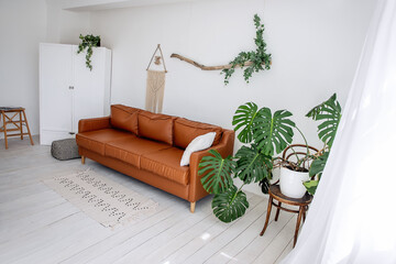 Stylish, trendy interior in Scandinavian style. In the white loft room there is brown leather sofa, green monster, driftwood with ivy hangs on the wall, handmade hammock hangs, garland of light bulbs