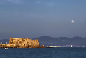 Landscape at sunset of Castel dell' Ovo, Naples with the turquoise sea, a sailboat and the moon rising behind the mountains