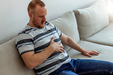 Young man suffering from stomach ache, sitting on a sofa in the living room. Man holding stomach in...