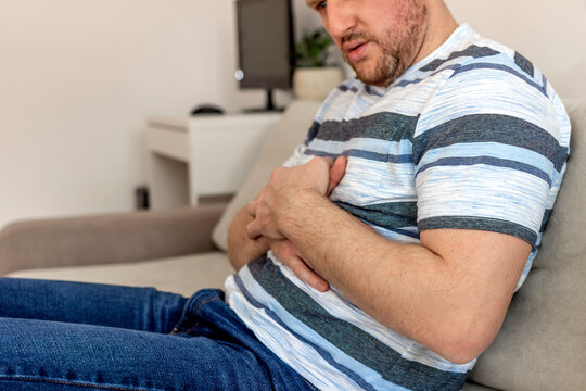 Serious mature man wearing casual clothing sitting on couch at home with hand on stomach cause nausea, painful disease feeling unwell. Caucasian male suffering from stomach problems. Copy space.