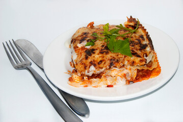 Portion of lasagna bolognese on white plate and covered with knife and fork on white background
