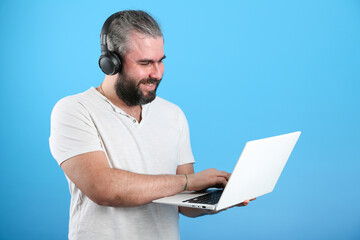 bearded man in headphones smiles and looks into a laptop on blue background. the concept of communicating with friends online via the internet 