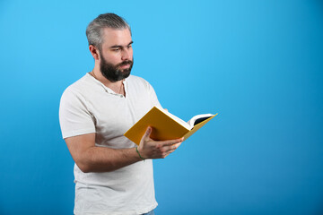 bearded man attentively reads a book on a blue background. 35-year-old man holding an open book 