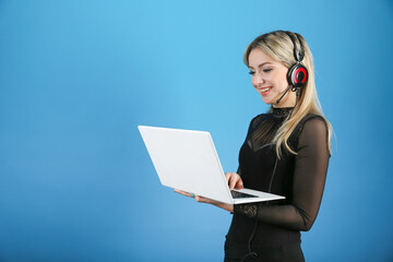 young woman wearing headphones with a microphone holds a laptop on a blue background. female dispatcher smiling over blue background. positive online meeting concept  