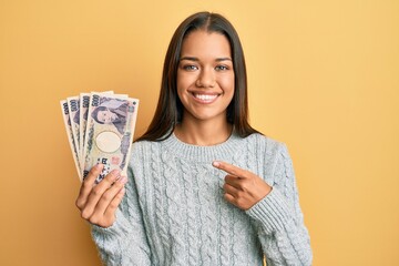 Beautiful hispanic woman holding 5000 japanese yen banknotes smiling happy pointing with hand and finger