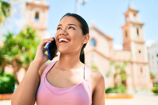 Young beautiful hispanic sporty woman wearing fitness outfit smiling happy and natural calling using smartphone at the town