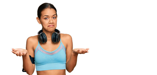Beautiful hispanic woman wearing sportswear and headphones clueless and confused expression with...