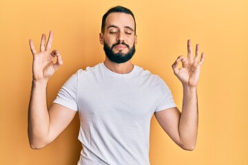 Young man with beard wearing casual white t shirt relax and smiling with eyes closed doing meditation gesture with fingers. yoga concept.