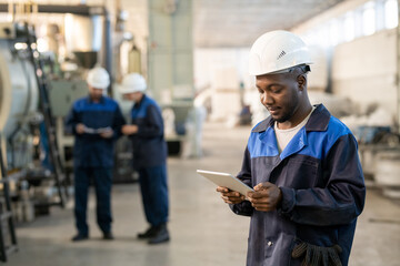 Young African male factory worker in overalls and hardhat using digital tablet while standing in...