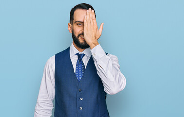 Young man with beard wearing business vest covering one eye with hand, confident smile on face and surprise emotion.