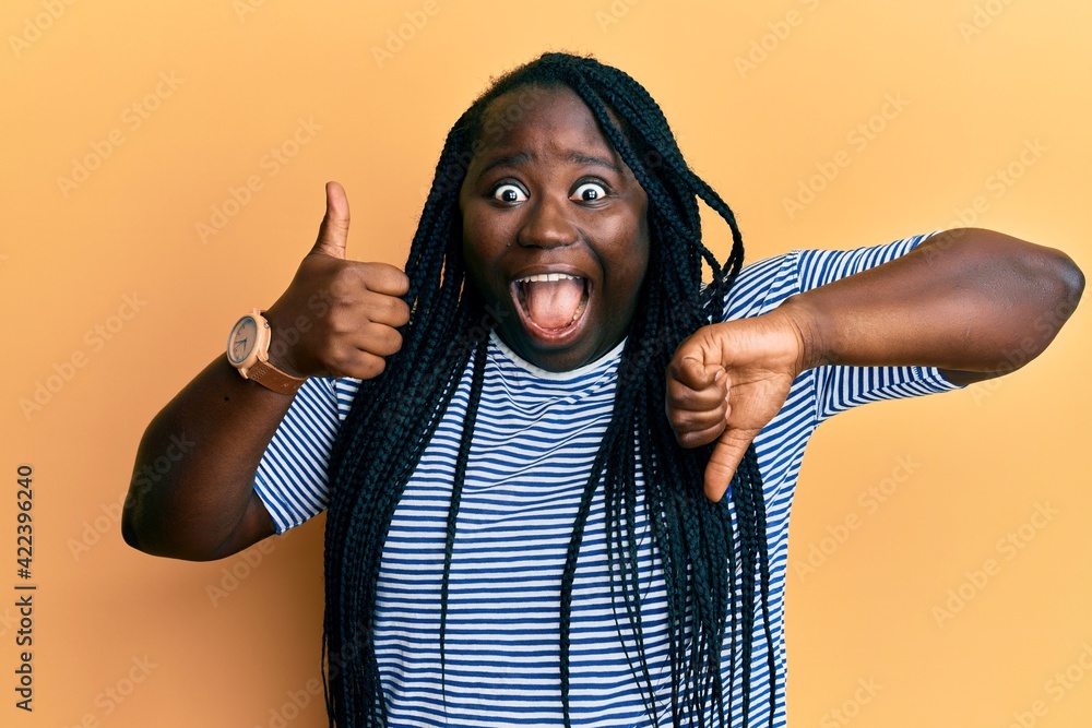 Sticker Young black woman with braids doing thumbs down and thumbs up gesture celebrating crazy and amazed for success with open eyes screaming excited.