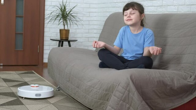 Meditation with a robot. A little girl in a lotus position meditates on the couch, a robot vacuum cleaner cleans the apartment.