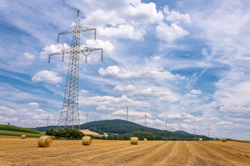 Strommast einer Hochspannungsleitung auf Stoppelfeld mit Strohballen und Windkraftanlagen