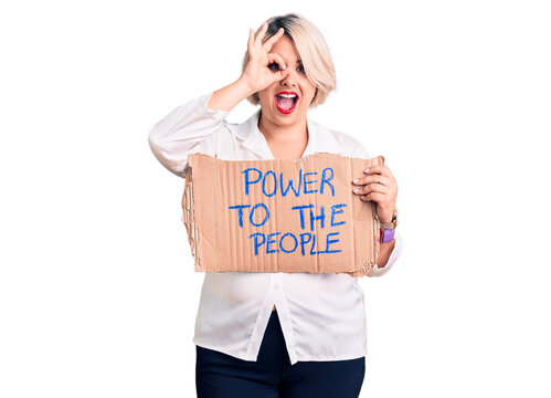 Young blonde plus size woman holding power to the people banner smiling happy doing ok sign with hand on eye looking through fingers