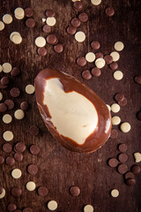 Top view of a dark and white chocolate easter egg with lots of chocolate chips all around on a wooden table.