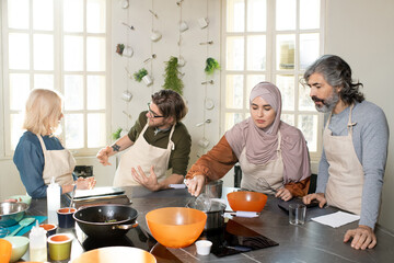 Young Muslim female trainee in hijab holding whisker over pan with water on electric stove while cooking at masterclass among other learners