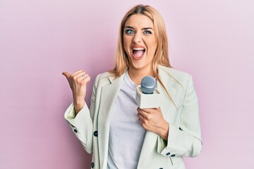 Young caucasian woman holding reporter microphone pointing thumb up to the side smiling happy with...