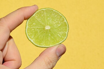 Slice of fresh lime fruit held in fingers of left hand of adult man, yellow wall in background. 