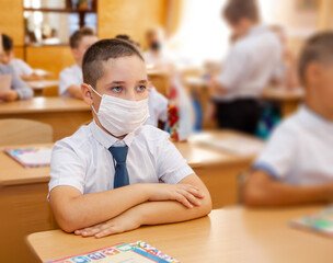 Beautiful schoolboy sitting in class. Boy doing schoolwork at classroom desks. Group of school kids with face masks writing test. Education, learning, schooling during quarantine. New normal
