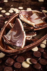 Dark and white chocolate easter egg with cracked eggs inside and lots of chocolate chips all around on a wooden table.