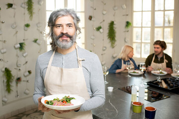 Mature bearded man in pullover and apron holding plate with selfmade vegetable salad and meat while standing against two people eating lunch