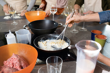 Hands of mature woman mixing up milk and butter in frying pan against other trainees cooking new course by large table in the kitchen