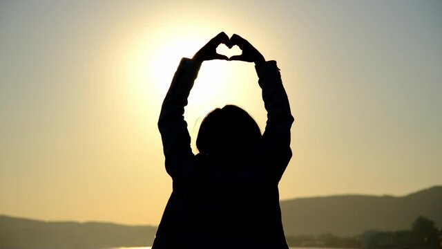 A young woman makes a heart sign by putting her hands together on the beach at sunset.