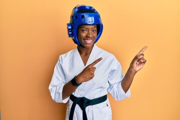 Young african american girl wearing taekwondo kimono and protection helmet smiling and looking at the camera pointing with two hands and fingers to the side.