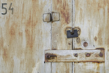 A rectangular padlock hangs from the old rusty metal doors of an abandoned garage or warehouse next to a large deadbolt and number 54. Storage of old things