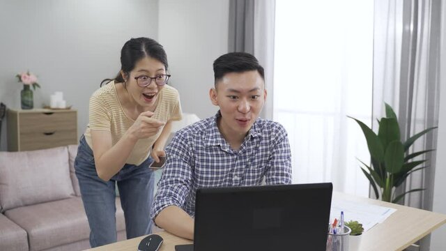 japanese woman is pointing at the screen and jumping for joy as her boyfriend is telling her to come and check out the job offer letter he’s got.
