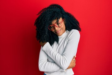 African american woman with afro hair wearing casual sweater and glasses hugging oneself happy and...