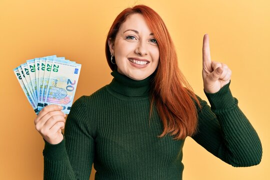 Beautiful redhead woman holding bunch of 20 euro banknotes smiling with an idea or question pointing finger with happy face, number one