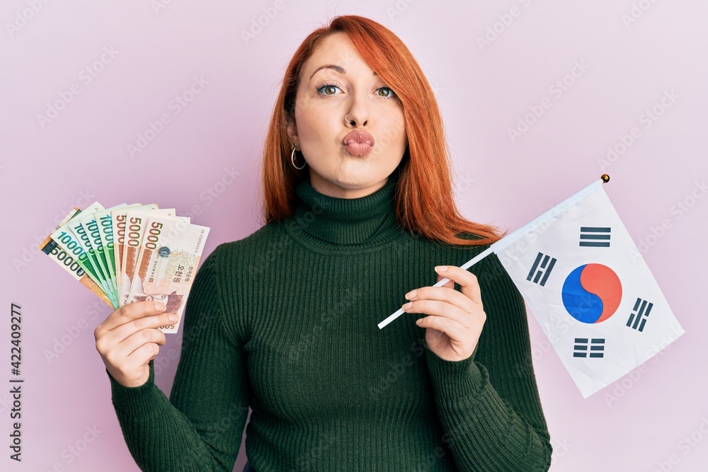 Sticker Beautiful redhead woman holding south korean won banknotes and flag looking at the camera blowing a kiss being lovely and sexy. love expression.