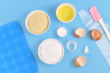 Ingredients for making homemade French Macarons sweets including powdered sugar, ground almonds, egg white, salt and baking tools like mold mat and icing pipe on blue background