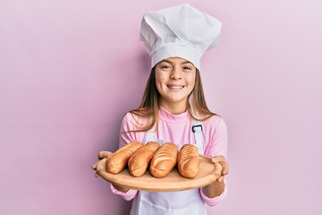 Beautiful brunette little girl wearing baker uniform holding homemade bread smiling and laughing hard out loud because funny crazy joke.