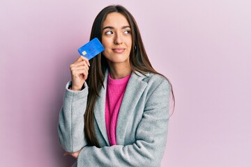 Young hispanic girl holding credit card smiling looking to the side and staring away thinking.