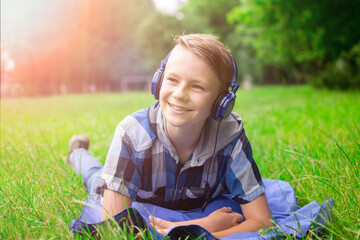 Laughing kid boy 12-16 year old with headphones outdoors. Childhood. Schoolboy. Lying on grass, summer.