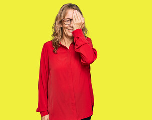 Middle age blonde woman wearing casual shirt over red background covering one eye with hand, confident smile on face and surprise emotion.